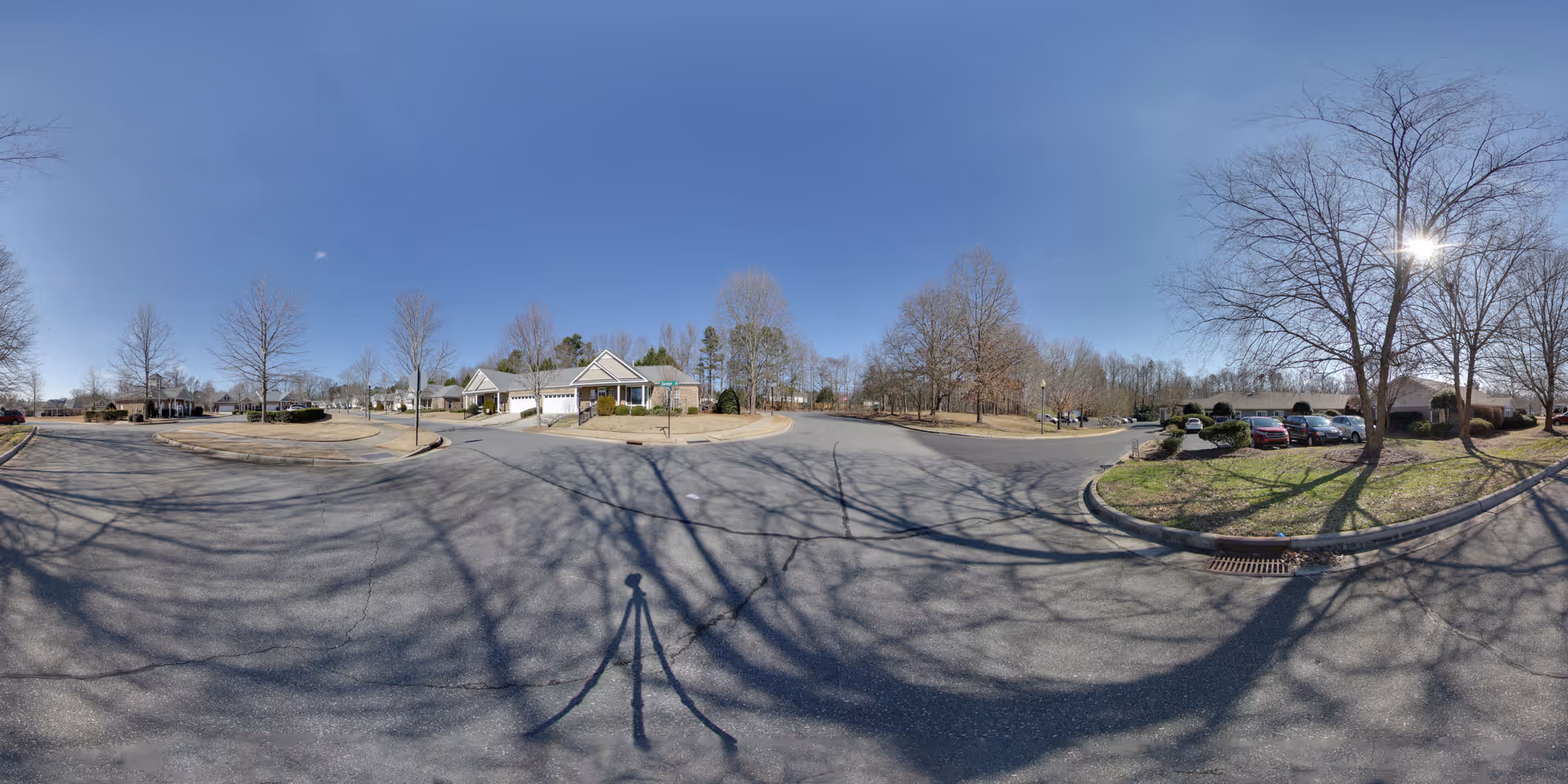 Wide panoramic view of a senior living facility parking area with leafless trees casting shadows on the asphalt. Several parked cars are visible on the right side near landscaped areas with grass and bushes. Residential-style buildings with garages are seen in the background under a clear blue sky.
