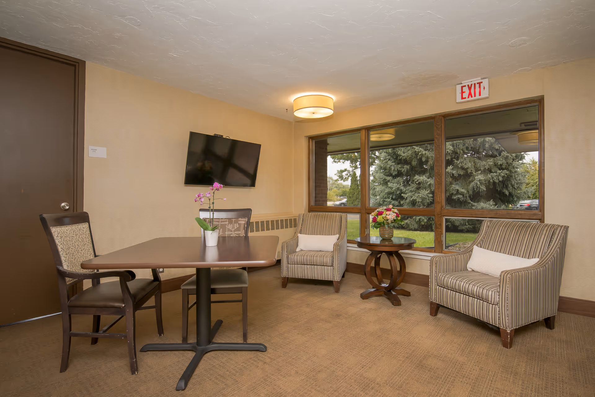 A cozy seating area with a table and chairs, two striped armchairs, a wall-mounted TV, and a large window showing trees outside.