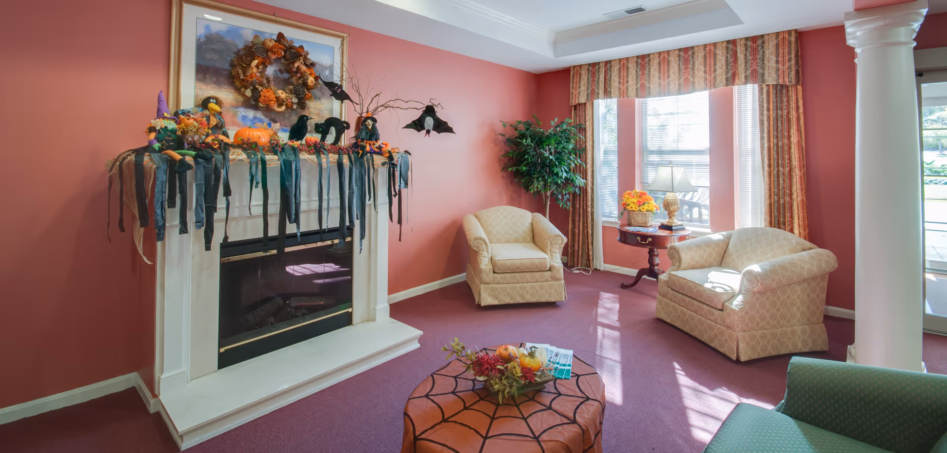 A cozy senior living room with a decorated white fireplace, upholstered armchairs, a coffee table and a window letting in sunlight.