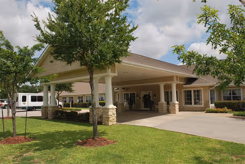 Covered front entrance of a single-story senior living building with a porte-cochere, lawn, and a parked white van.