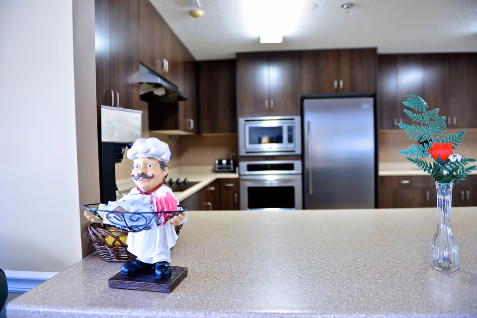 A modern kitchen with dark wooden cabinets, stainless steel appliances including a refrigerator, microwave, and oven. On the countertop in the foreground, there is a small chef figurine holding a basket with napkins and a glass vase with a red artificial flower and green leaves.