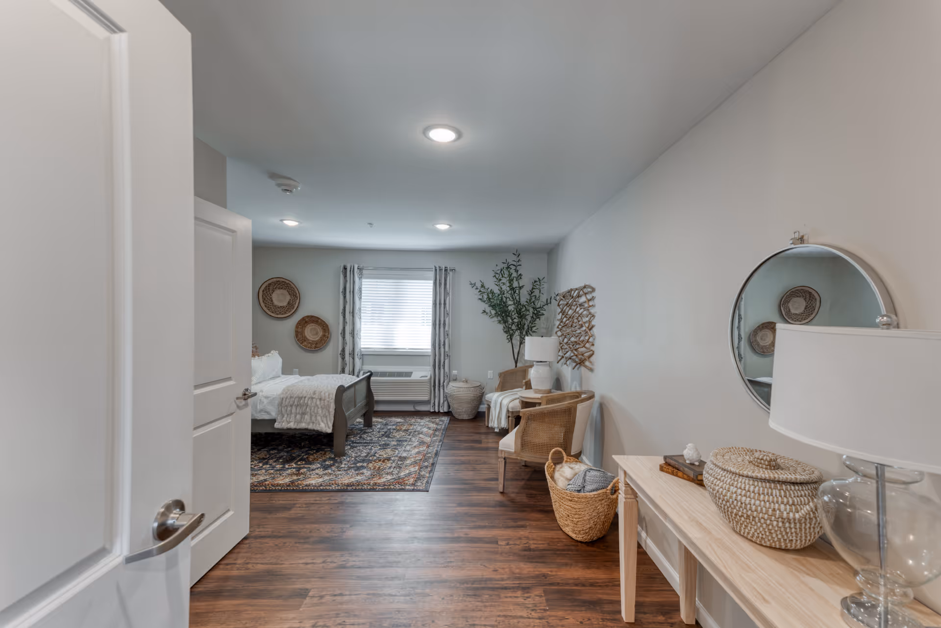 View into a senior living bedroom with a wooden bed, patterned area rug, two wicker chairs with a small table and lamp between them, a round mirror on the wall, decorative baskets, and a window with curtains letting in natural light.