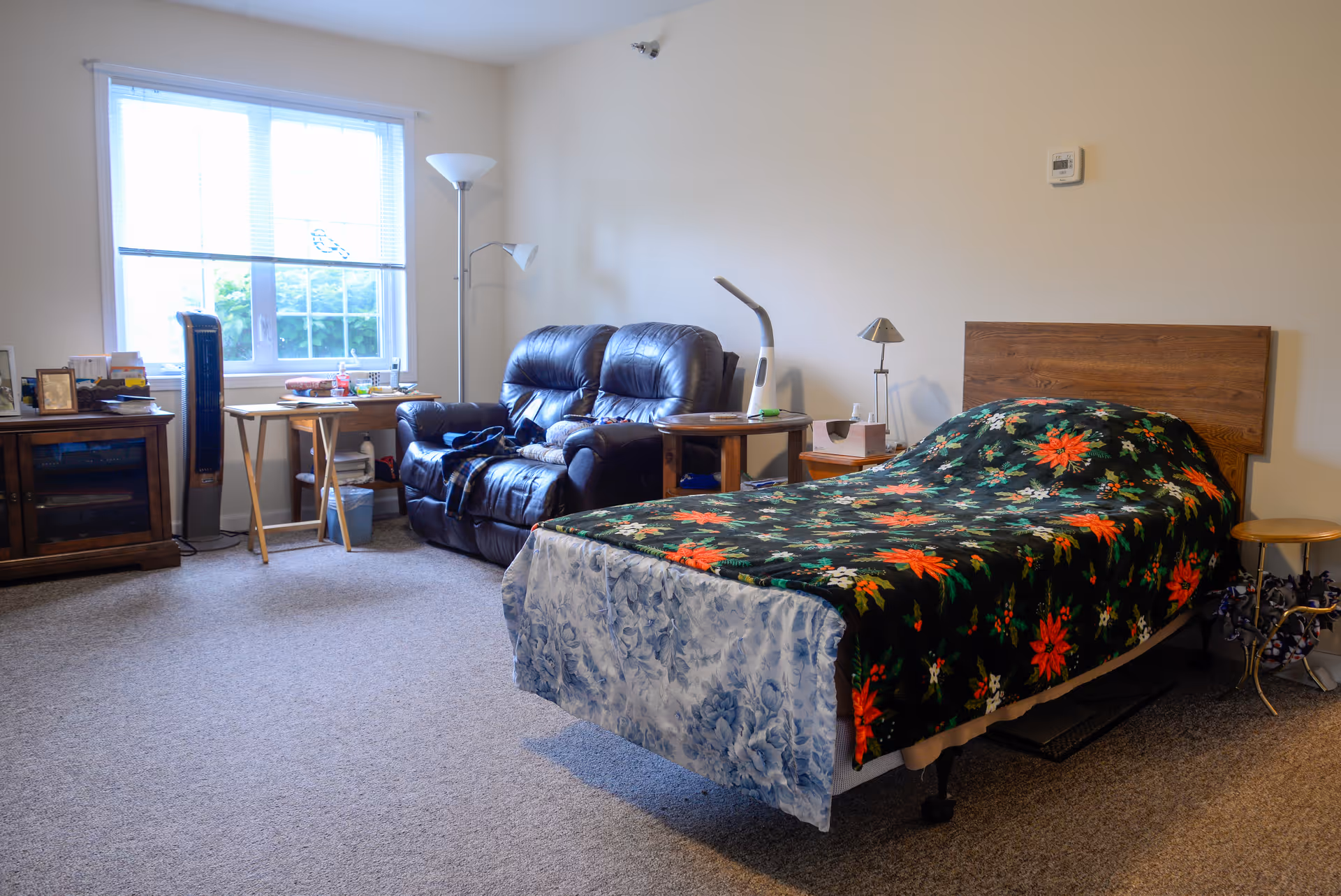 A cozy bedroom in a senior living facility with a single bed covered by a floral blanket, a dark leather loveseat, a wooden side table with a lamp, a small wooden table near a window with blinds, and a standing floor lamp. The room has beige walls and carpeted floor.