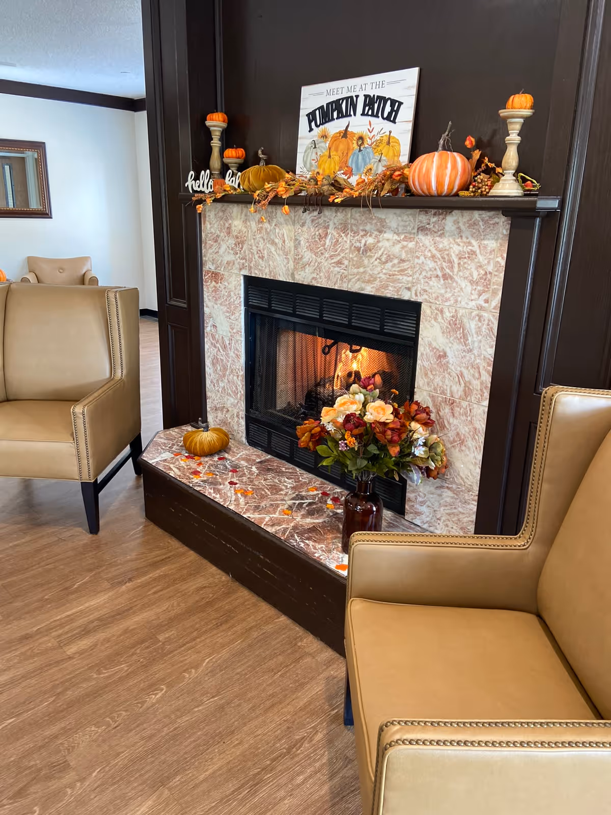 A cozy living room area featuring a lit fireplace with a marble surround and dark wood paneling. The mantel is decorated with autumn-themed items including small pumpkins, candles, and a sign that reads 'Meet me at the Pumpkin Patch.' There is a vase with a bouquet of flowers on the hearth, and two tan leather armchairs positioned nearby on a wood floor.