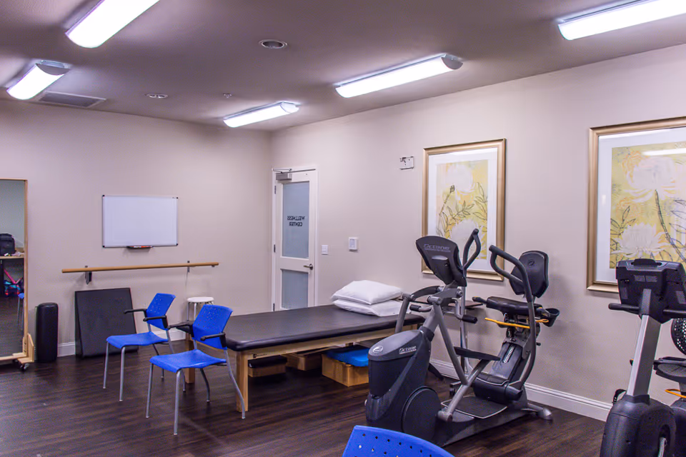 A wellness center room with exercise equipment including stationary bikes, a treatment table with pillows, two blue chairs, a whiteboard on the wall, and framed floral artwork. The room has wooden flooring and fluorescent ceiling lights.