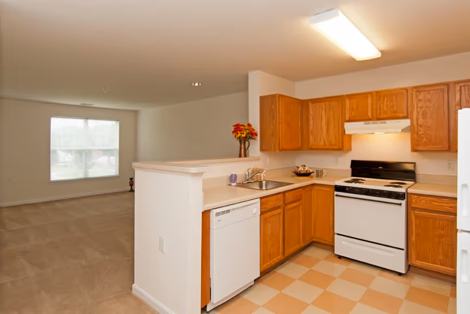 Interior view of a kitchen with wooden cabinets, a white stove, dishwasher, and refrigerator. The kitchen opens into a carpeted living area with a large window letting in natural light.