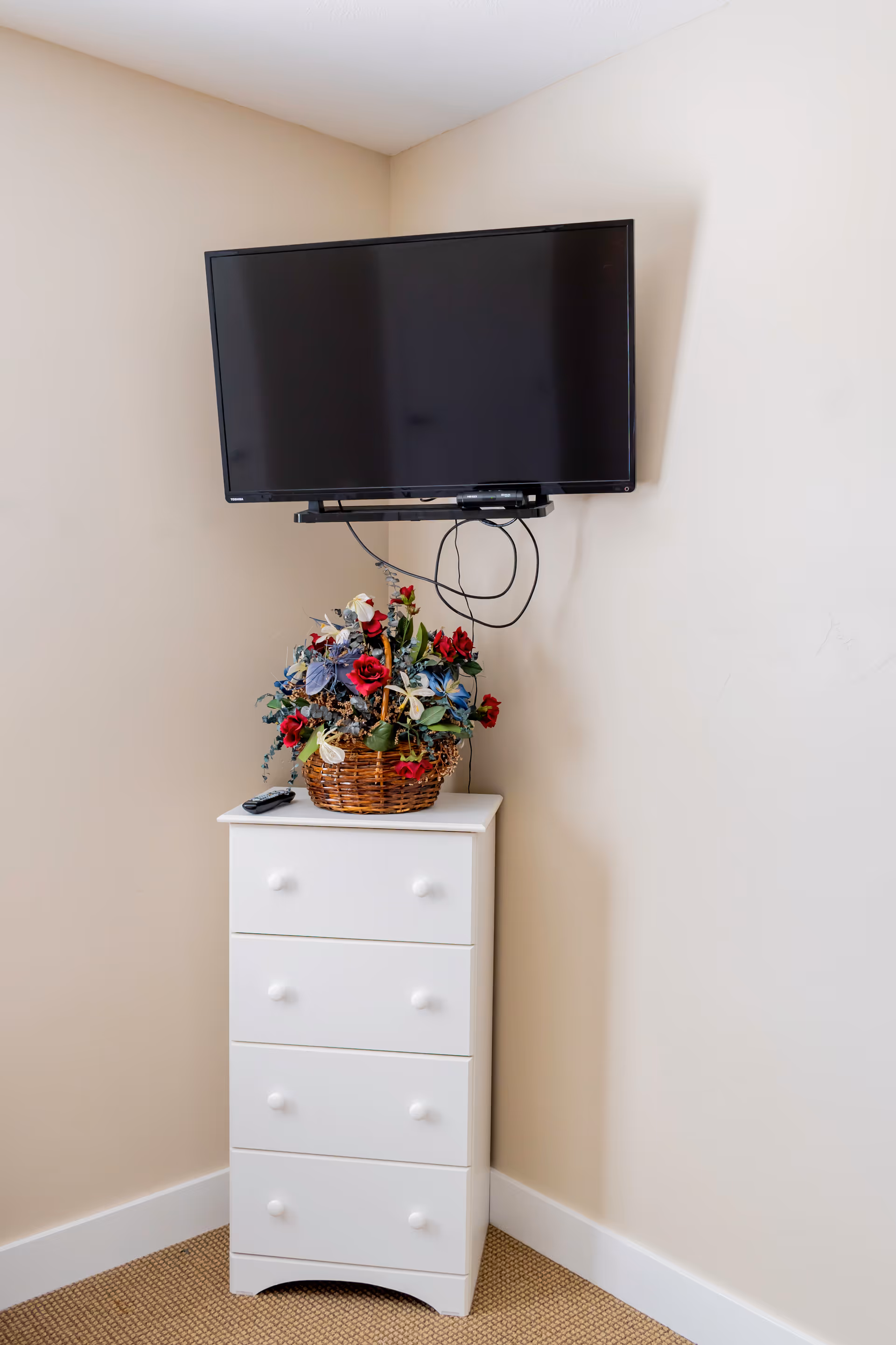 A corner of a room with a wall-mounted flat-screen TV above a white four-drawer dresser. On top of the dresser is a basket filled with artificial flowers in red, blue, and green colors. A TV remote is also placed on the dresser.
