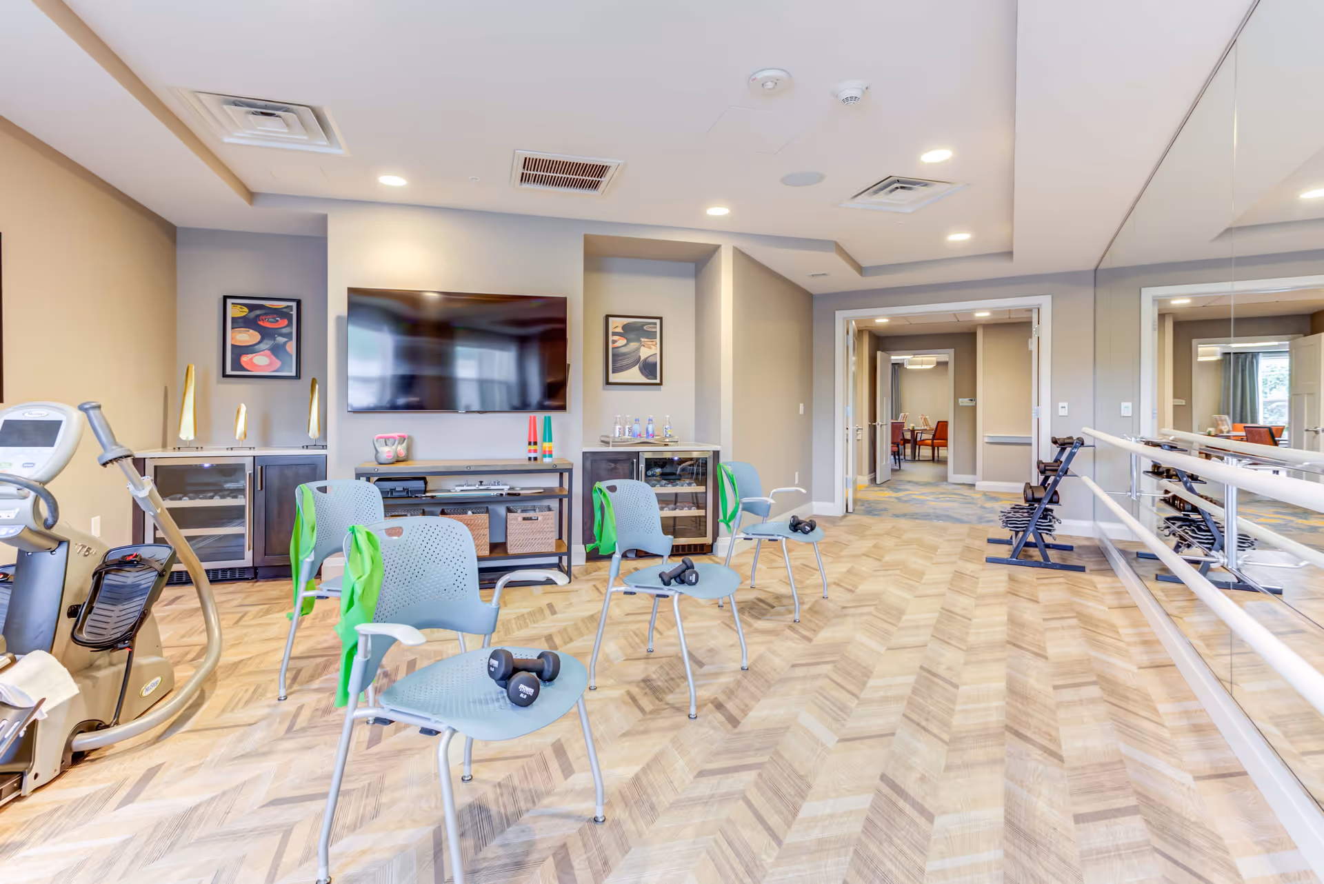 A fitness room in a senior living facility with exercise chairs equipped with dumbbells and resistance bands, a stationary exercise bike, a large wall-mounted TV, mirrored wall with ballet bars, and wooden flooring. The room is well-lit with recessed ceiling lights and has a neutral color scheme with beige walls and light wood patterned floor.