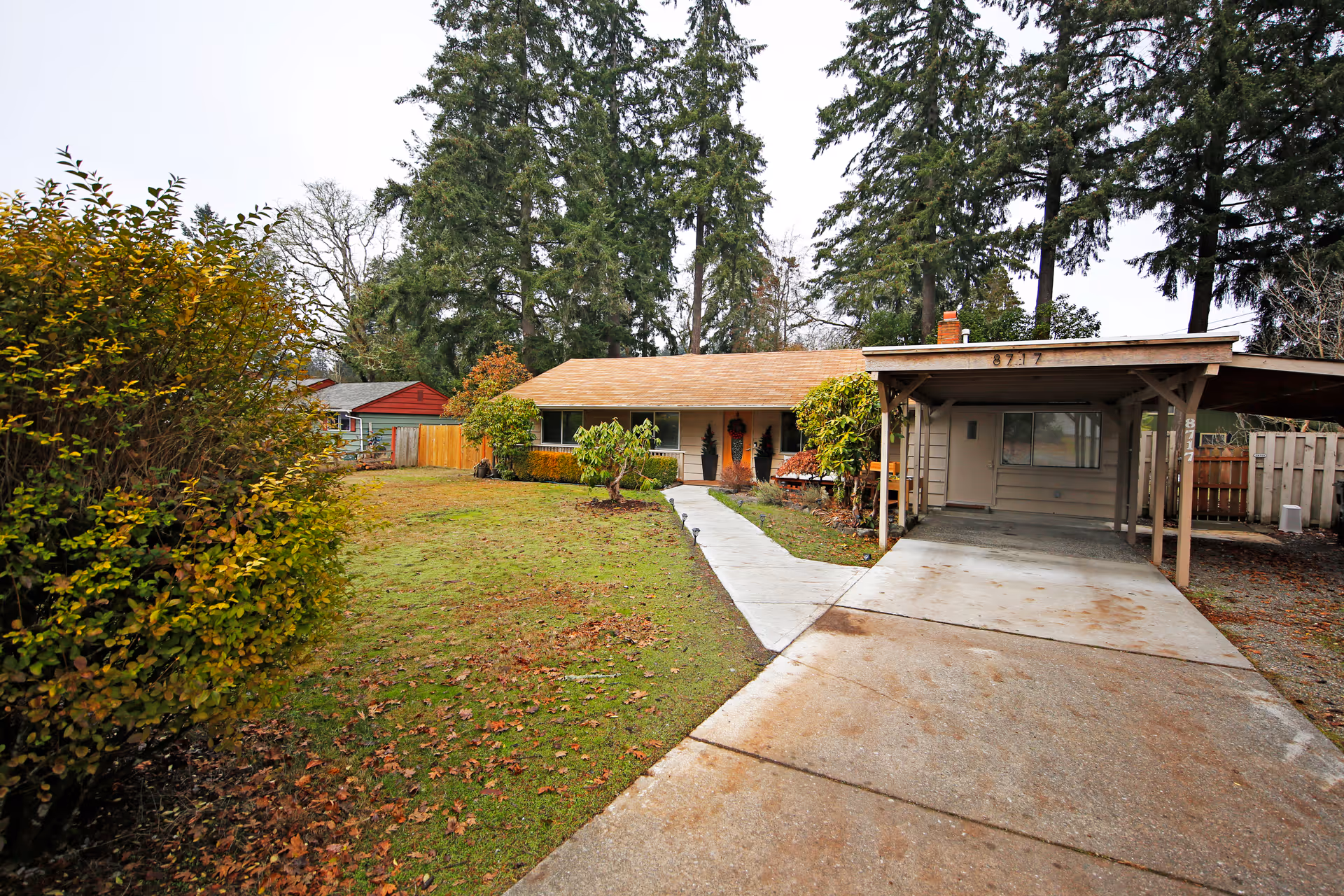 Front view of a single-story house with a carport, concrete driveway and walkway, and a grassy front yard with trees.