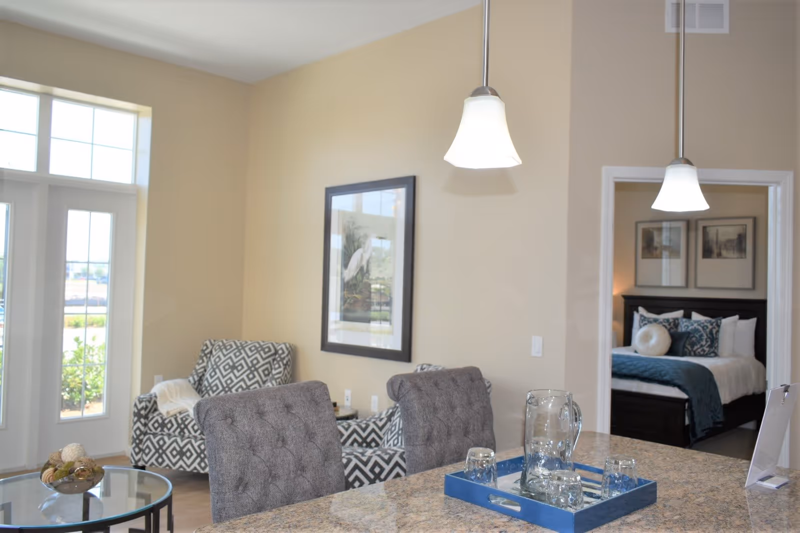 Interior view of a senior living facility showing a granite countertop with a blue tray holding a glass pitcher and four glasses. Two gray upholstered chairs are positioned at the counter. In the background, there are two patterned armchairs near large windows with natural light coming in. A bedroom with a neatly made bed and framed pictures on the wall is visible through an open doorway. Two pendant lights hang from the ceiling.