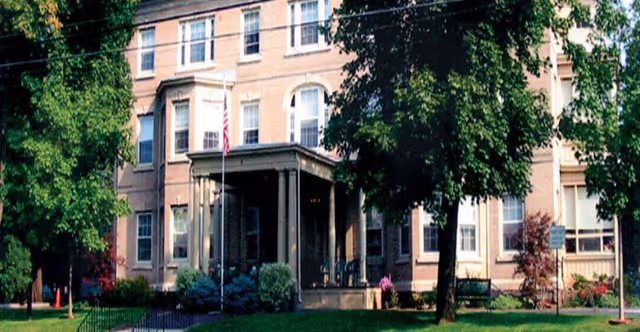 Exterior view of a multi-story brick building with large windows, a covered entrance with columns, surrounded by green trees and a well-maintained lawn. An American flag is displayed near the entrance.