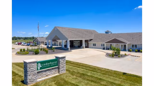 Exterior view of Cedarhurst Senior Living of Bethalto building with a large driveway, landscaped areas, and a sign in the foreground displaying the facility's name. The sky is clear and blue.