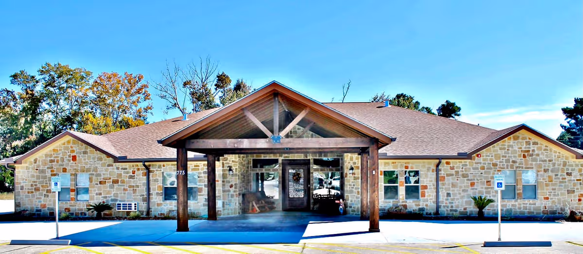 Front exterior view of a single-story assisted living facility building with a stone facade, a peaked roof, and a covered entrance supported by wooden beams. There are two handicap parking signs visible in front of the building, and trees in the background under a clear blue sky.