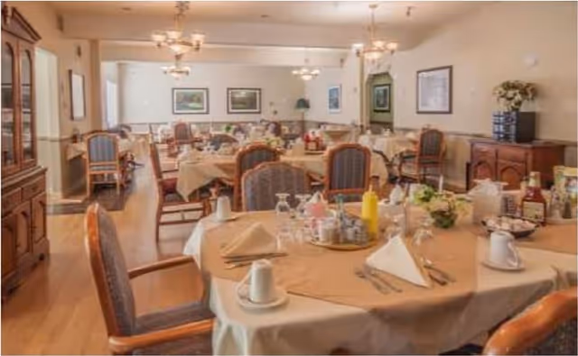 A dining room with multiple tables covered in beige tablecloths, set with napkins, cups, glasses, and condiments. The room has wooden chairs with cushioned seats and backs, wooden flooring, and framed artwork on the walls. There are chandeliers hanging from the ceiling providing warm lighting.
