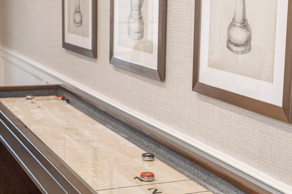 A shuffleboard table with pucks on it is positioned against a wall with three framed black and white sketches of chess pieces hanging above it.