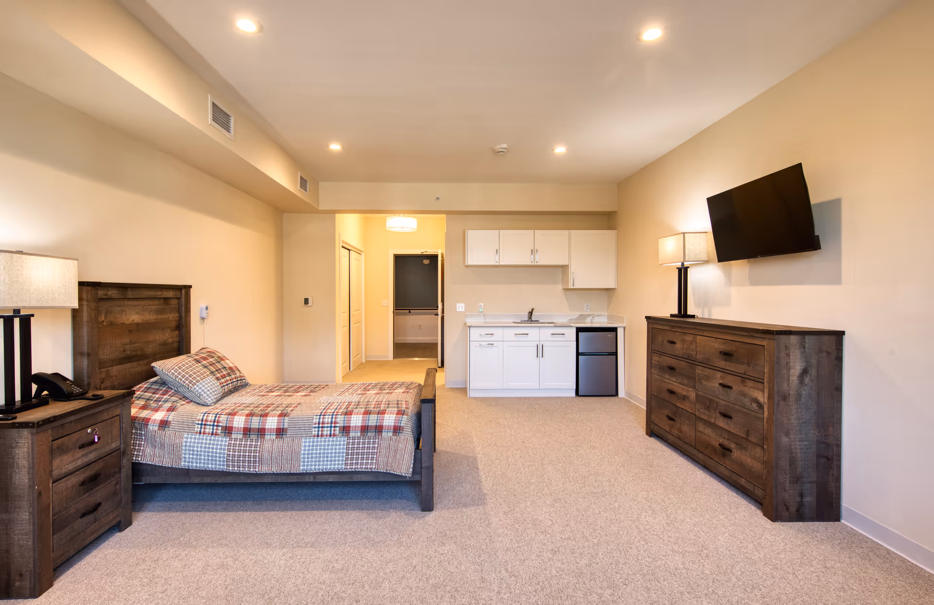 A spacious senior living studio room with a bed, bedside table, kitchenette, dresser, and wall-mounted TV.