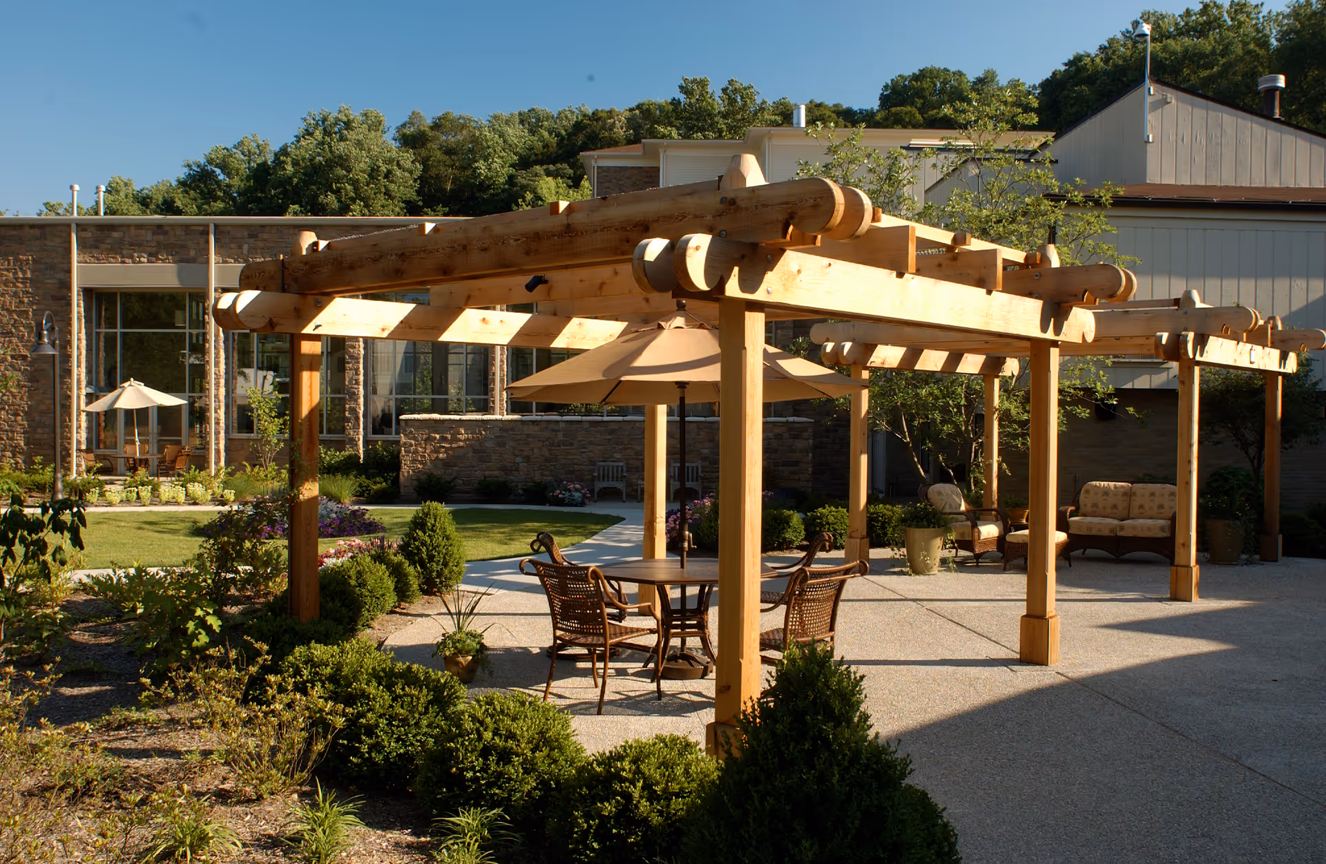 Outdoor patio area at Deupree House with wooden pergolas, round tables with umbrellas, wicker chairs, and cushioned seating. The area is surrounded by greenery, shrubs, and trees with a building in the background under a clear blue sky.