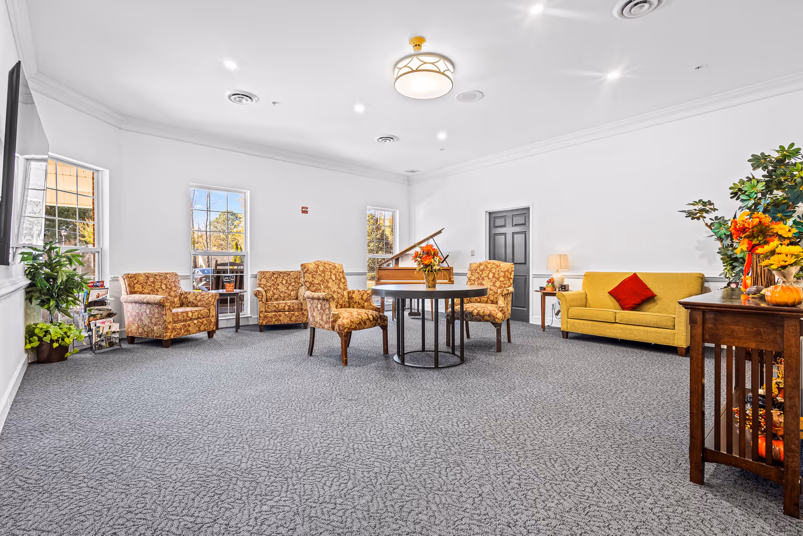 Bright communal living room with upholstered chairs, a yellow sofa, round table, piano, and decorative plants.
