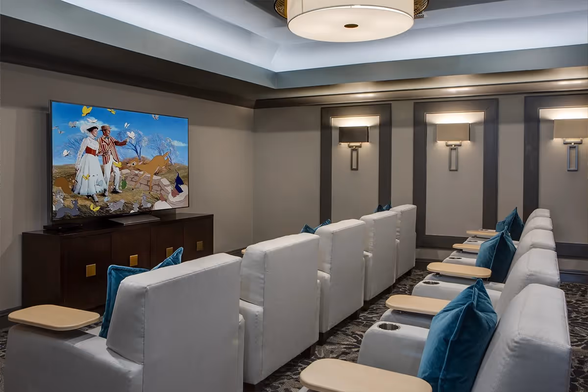 A cozy media room with two rows of white cushioned recliner chairs, each equipped with a small wooden tray and blue accent pillows. A large flat-screen TV is mounted on a dark wooden cabinet, displaying a colorful scene from a movie. The room features soft wall lighting and a patterned carpet.
