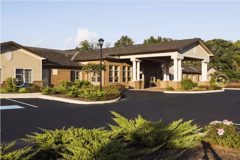Exterior view of Central Parke Assisted Living & Memory Care building with a covered entrance, brick and beige siding, surrounded by landscaped greenery and a paved driveway.