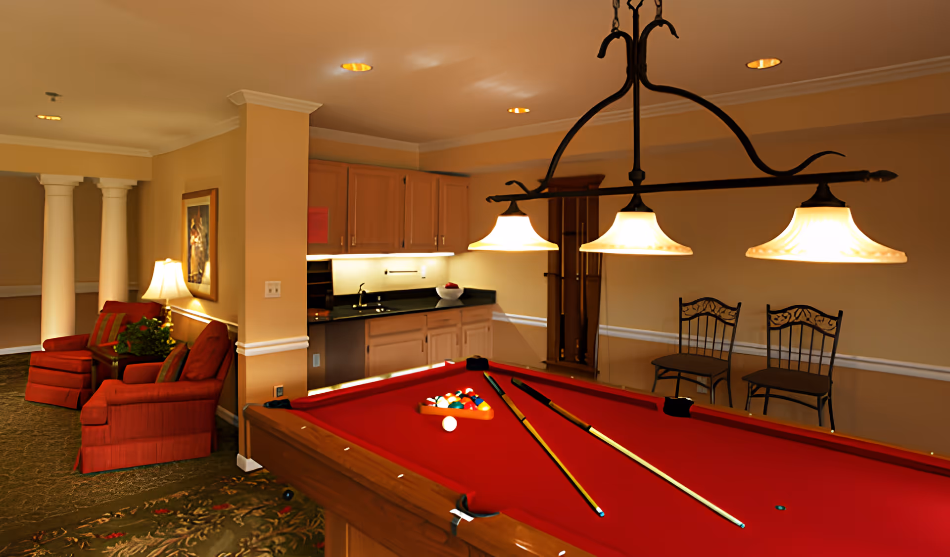 Interior of a recreational room featuring a red pool table with billiard balls and cues, overhead lighting with three lamps, two metal chairs against the wall, a small kitchenette with cabinets and a countertop, and a seating area with two red armchairs and a lamp near decorative white columns.