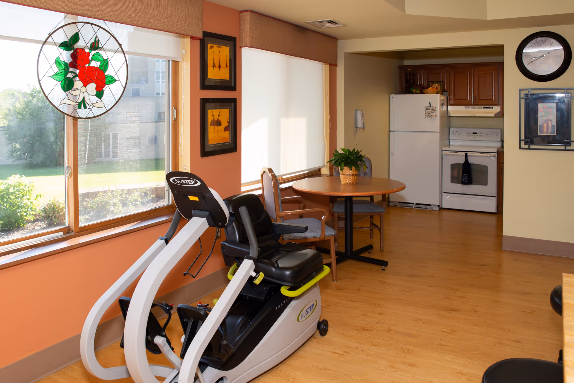 A bright room with large windows featuring a stained glass decoration, a recumbent exercise bike, a round wooden table with chairs, and a kitchen area with a refrigerator, stove, and cabinets. The room has wooden flooring and light-colored walls with some framed artwork and a clock on the wall.