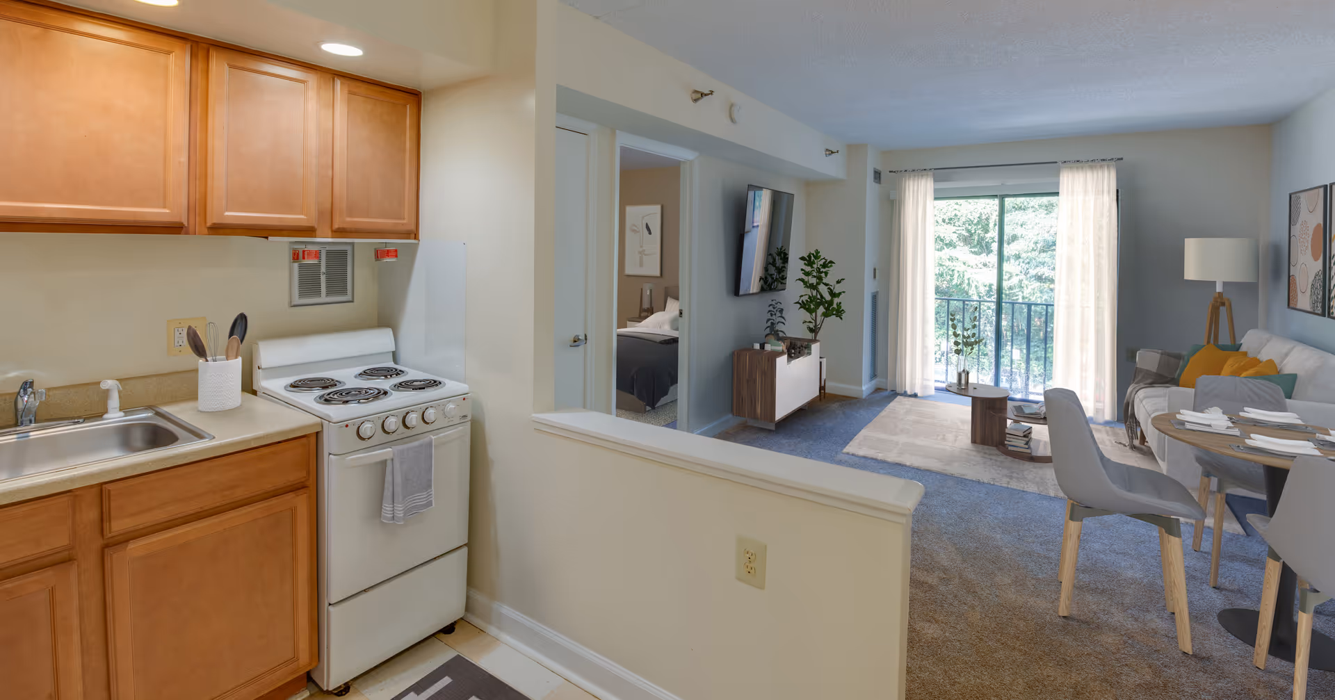 View of a small kitchen with wooden cabinets, a white electric stove, and a sink on the left side. The kitchen opens into a living room area with a wall-mounted TV, a plant, a sofa with colorful pillows, a floor lamp, and a dining table with chairs. A sliding glass door with sheer curtains leads to a balcony with greenery outside. A bedroom is partially visible through an open door in the background.
