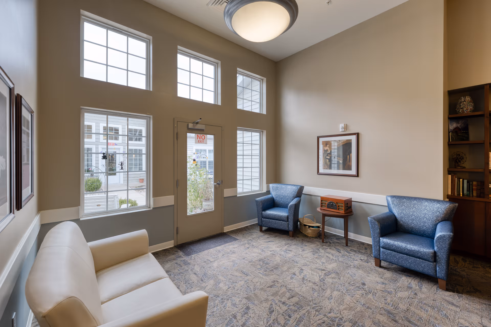 A cozy sitting area in a senior living facility with a beige couch on the left, two blue armchairs on the right, a small wooden table with a vintage radio between the armchairs, and large windows letting in natural light. The walls are painted beige and gray, and there is a framed picture on the wall above the armchairs.
