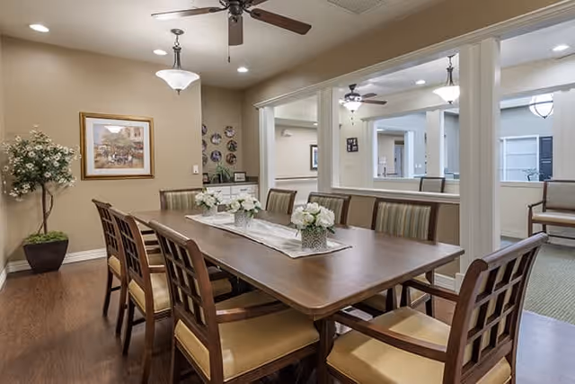 A well-lit dining room with a long wooden table surrounded by eight chairs with cushioned seats. The table is decorated with three small flower arrangements on a white runner. The room features beige walls, a ceiling fan with lights, framed artwork, and a potted plant in the corner. Large windows open to an adjacent room with additional seating.