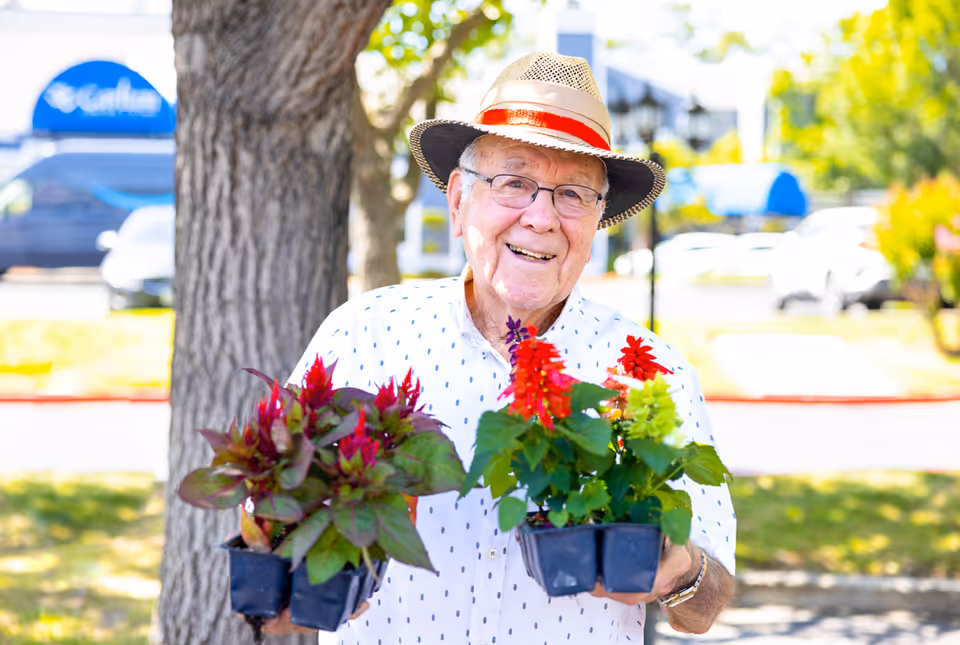 An elderly man wearing a straw hat with a red band and glasses is smiling while holding two small pots of colorful flowering plants outdoors, with trees and parked cars in the background.