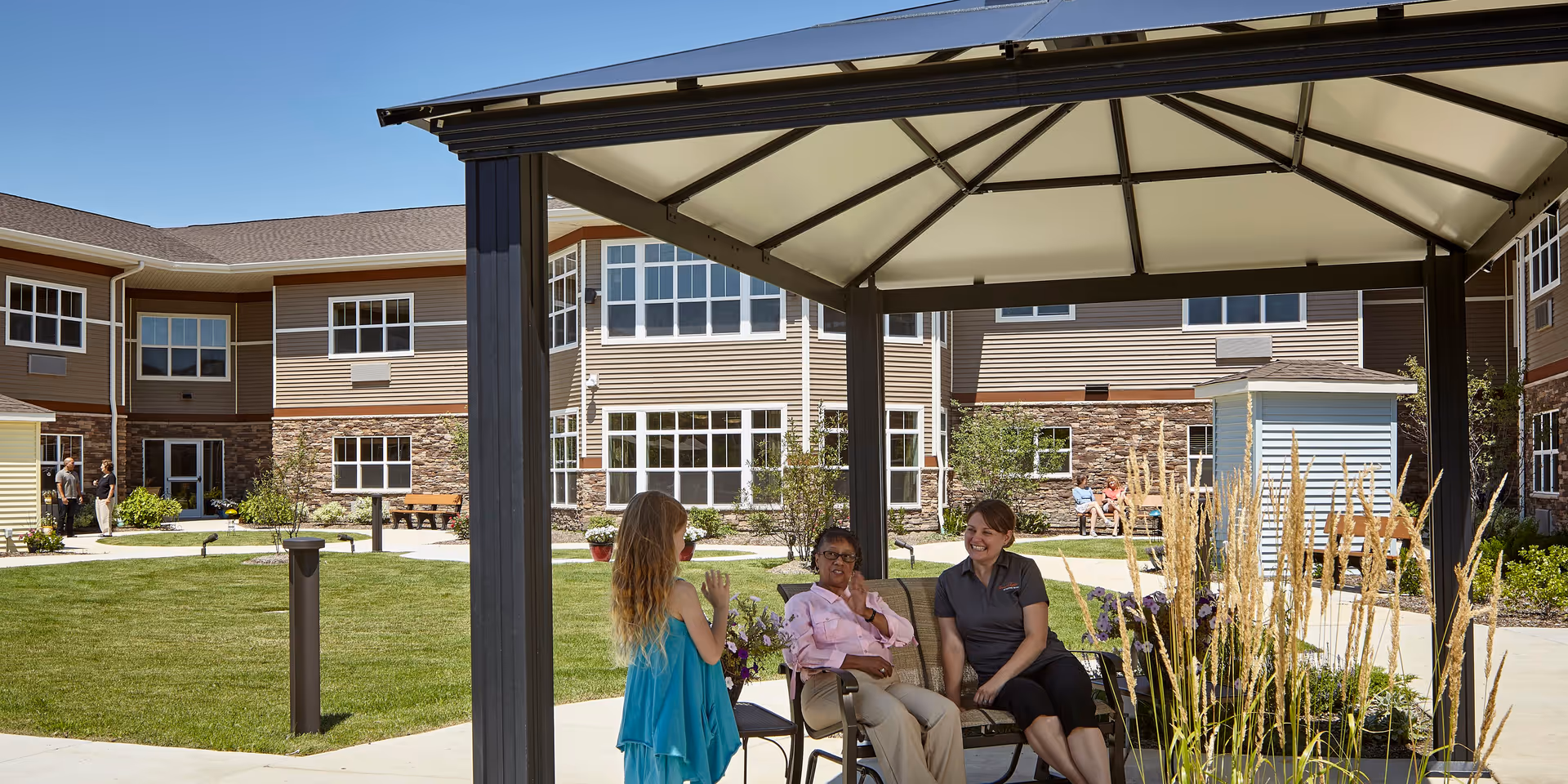 Two women sitting and chatting under a covered outdoor gazebo in a courtyard of a senior living facility, with a young girl standing nearby. The courtyard has green grass, plants, and a multi-story building with many windows in the background.