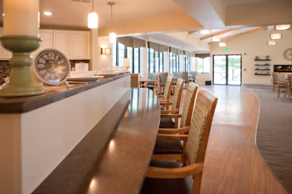 Interior dining area with a curved serving counter, lined chairs, tables, and large windows in a senior living facility.