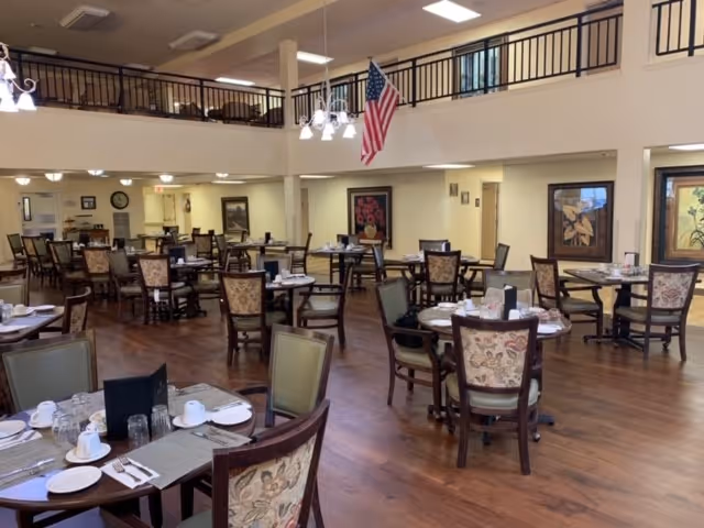 A spacious dining room with multiple round tables set with placemats, cups, glasses, and silverware. The room has wooden flooring, an American flag hanging from the ceiling, and a second-floor balcony railing overlooking the area. The walls are decorated with framed artwork.