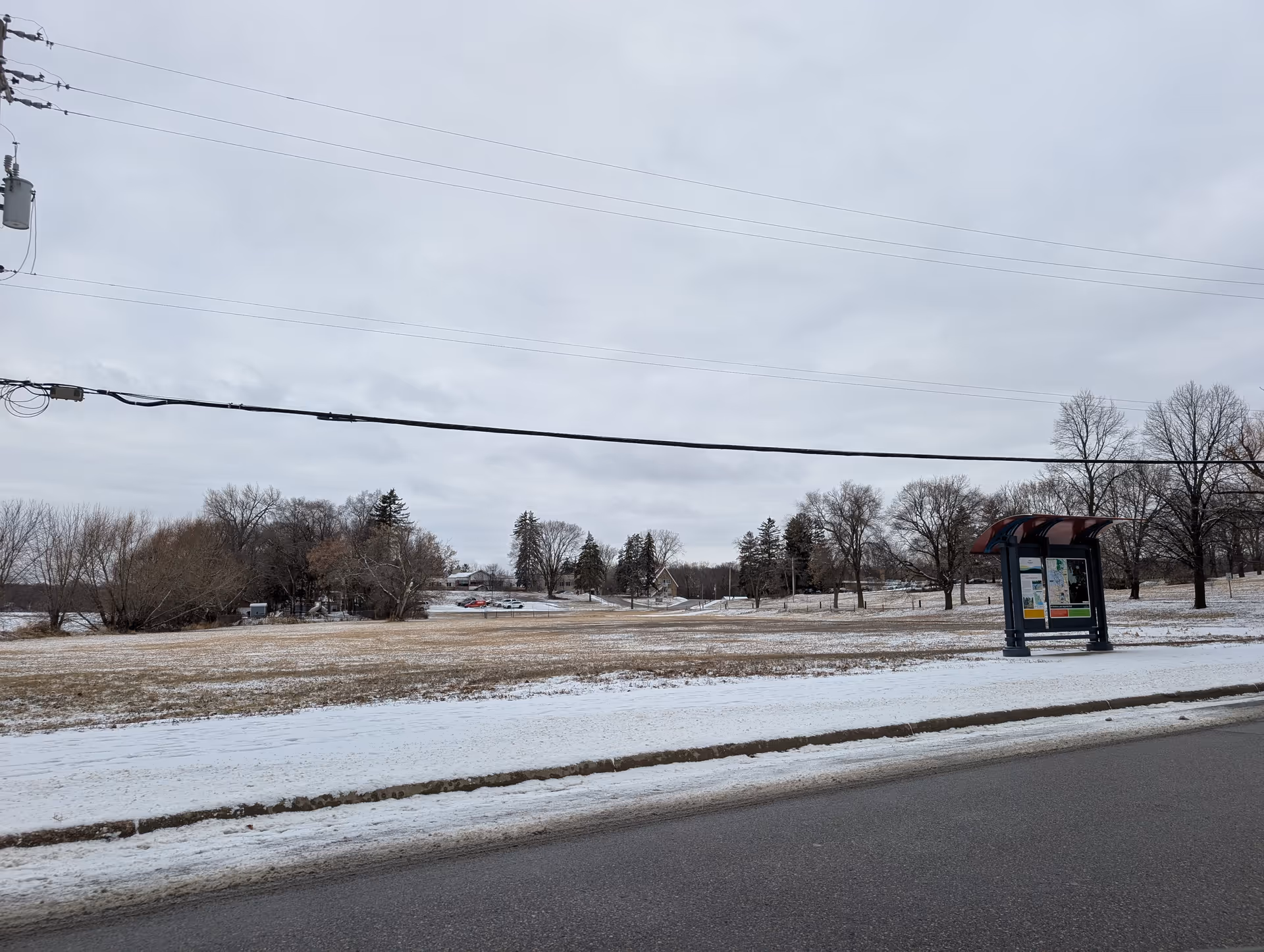 Snowy open field with scattered trees, a roadside bus shelter on the right, and overhead power lines.