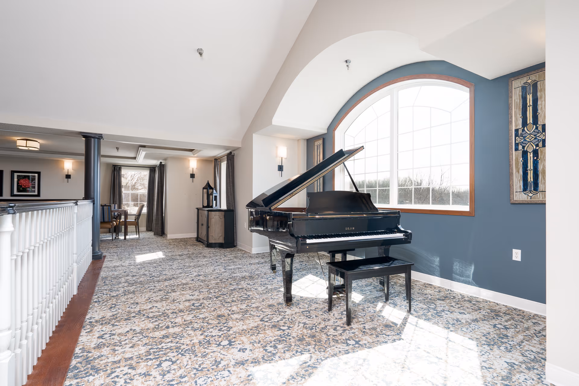 Sunlit interior common area with a black grand piano beside a large arched window and patterned carpet.