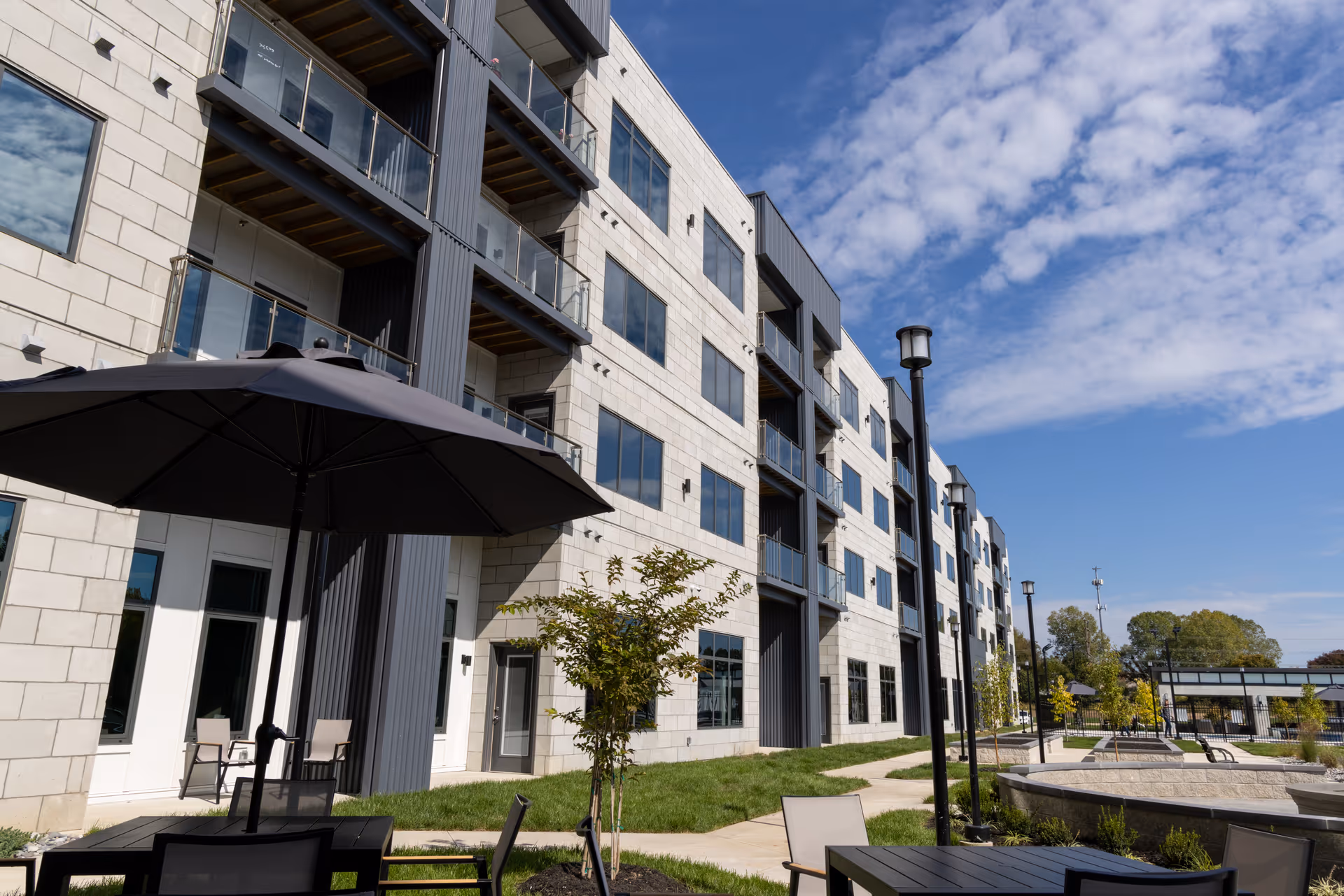 Modern multi-story senior living building with balconies and outdoor seating and umbrellas in a landscaped courtyard.