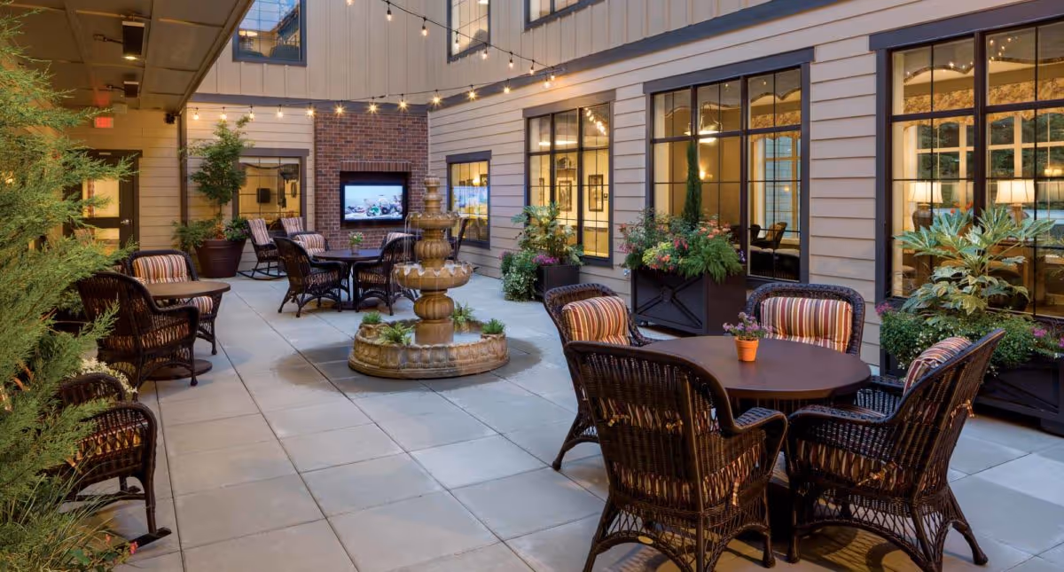 Outdoor courtyard area with several round tables surrounded by wicker chairs with striped cushions. There is a multi-tiered stone fountain in the center, potted plants along the walls, string lights overhead, and large windows showing the interior of the building.