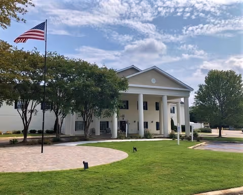 Front exterior view of a two-story building with white columns and a triangular pediment, surrounded by green grass, trees, and an American flag on a flagpole under a partly cloudy sky.