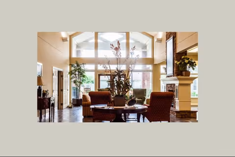 A bright and spacious living room area with large windows letting in natural light. The room features comfortable upholstered chairs arranged around a wooden coffee table with a large plant centerpiece. There is a fireplace on the right side with a framed picture above it and additional plants placed on the mantel. The walls are painted in warm tones, and the floor is wooden.