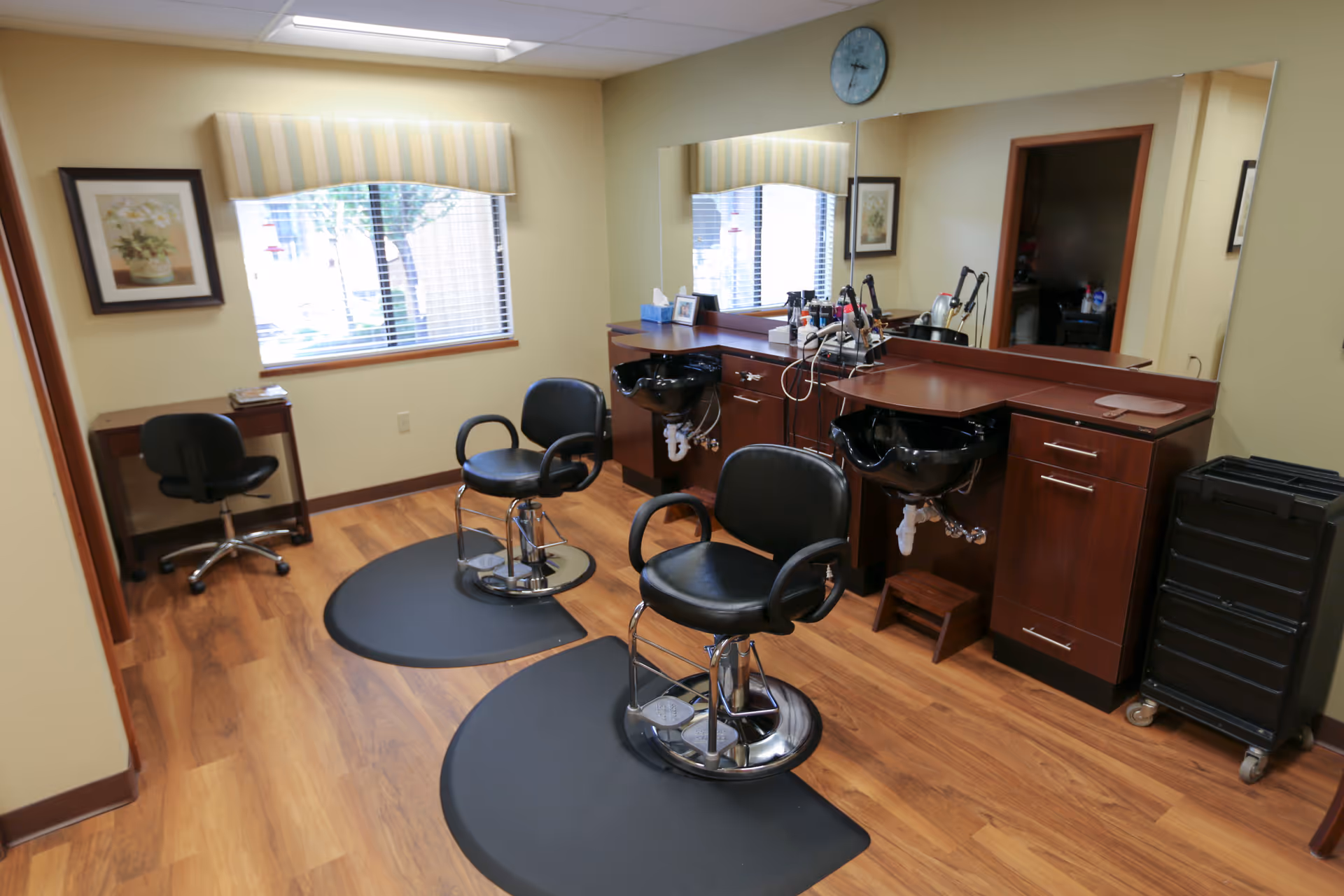 Interior view of a hair salon area in a senior living facility with two black salon chairs on black mats in front of a large mirror. There are two hair washing sinks with wooden cabinetry underneath, various hair styling tools on the counter, a clock on the wall, a window with blinds and a striped valance, a small desk with a chair, and framed artwork on the walls.