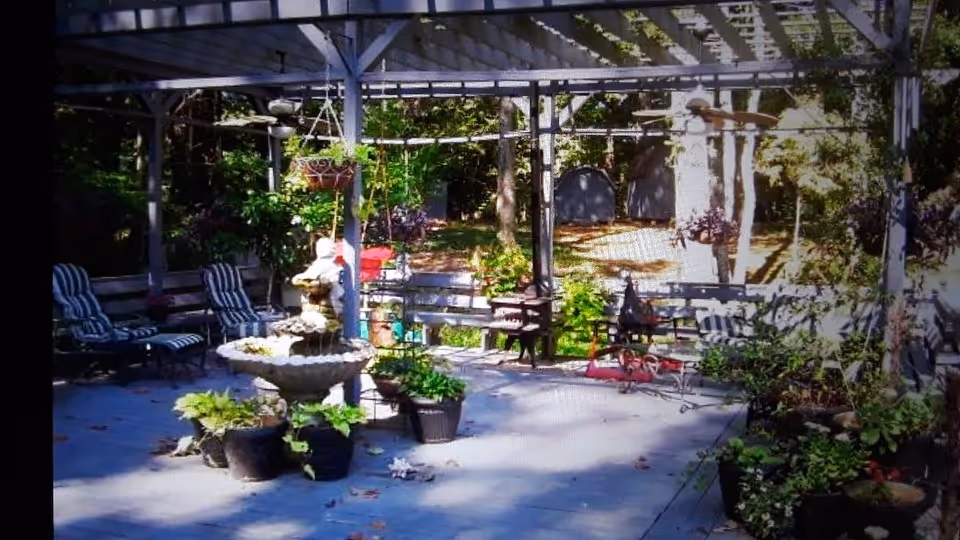 Outdoor patio area with a pergola overhead, featuring a central stone fountain surrounded by potted plants. Several cushioned lounge chairs and benches are arranged around the space, with hanging flower baskets and greenery in the background.
