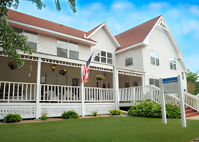A large white building with a red roof and a spacious covered porch featuring white railings. An American flag is hanging from the porch. There are green bushes and a well-maintained lawn in front of the building under a blue sky with some clouds.