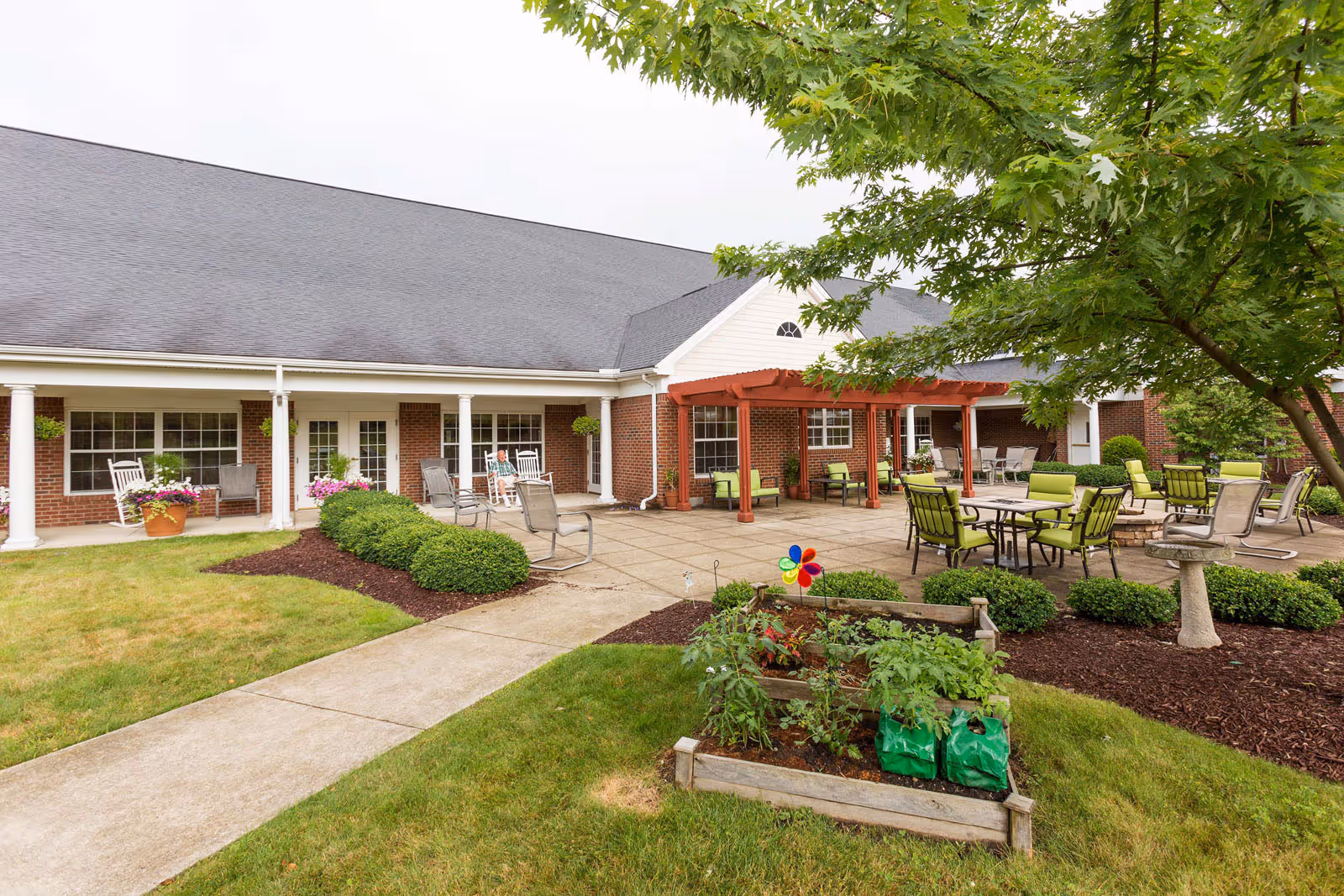 Outdoor patio area at Independence Village of Aurora featuring a paved seating area with green cushioned chairs and tables under a wooden pergola. There are bushes and a small garden bed with plants and a colorful pinwheel in the foreground. The building has a brick exterior with white columns and large windows. A person is sitting on a rocking chair near the building.