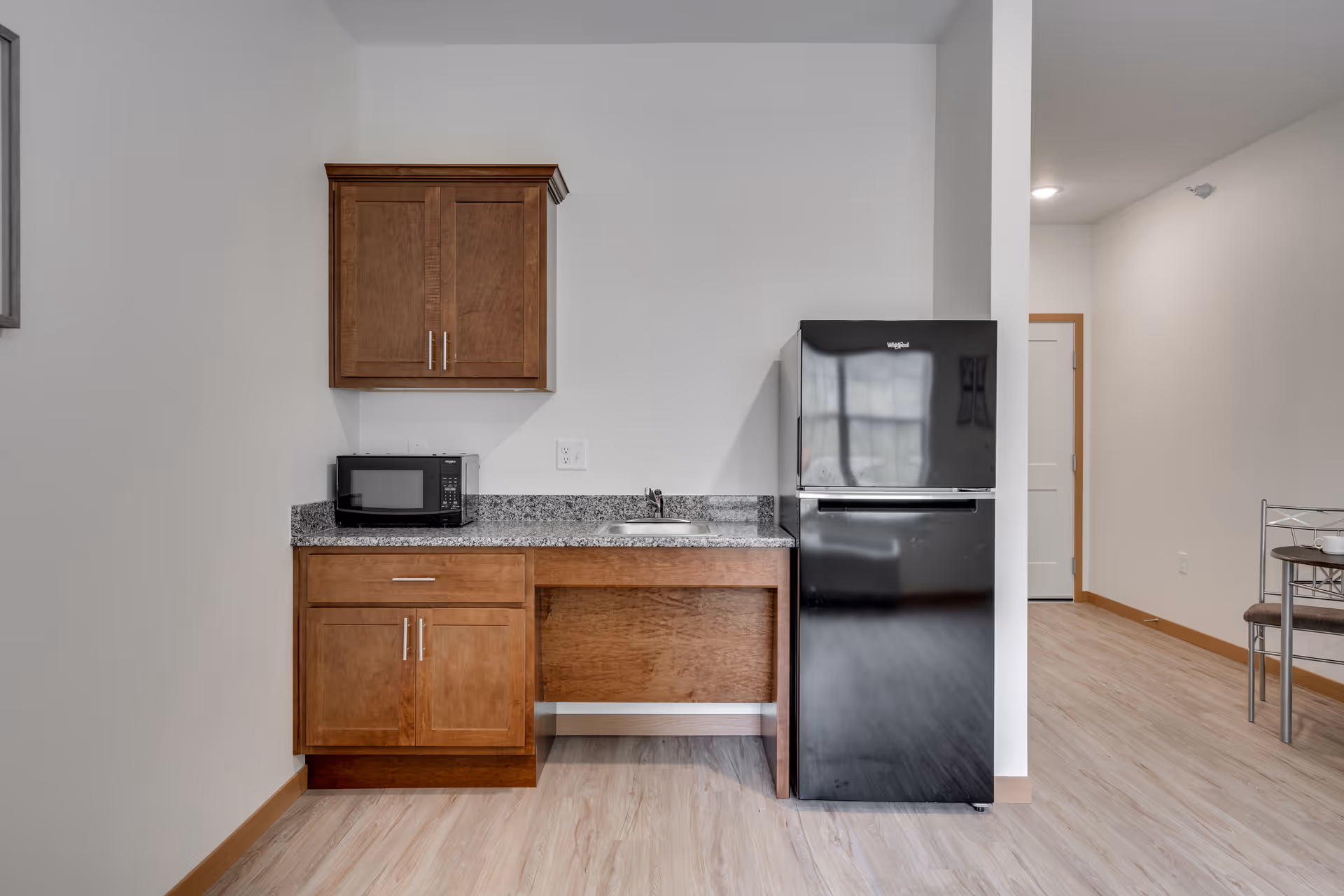 A small kitchen area with wooden cabinets, a granite countertop, a black microwave, a small sink, and a black Whirlpool refrigerator. The floor is light wood, and there is a partial view of a dining table and chairs to the right.
