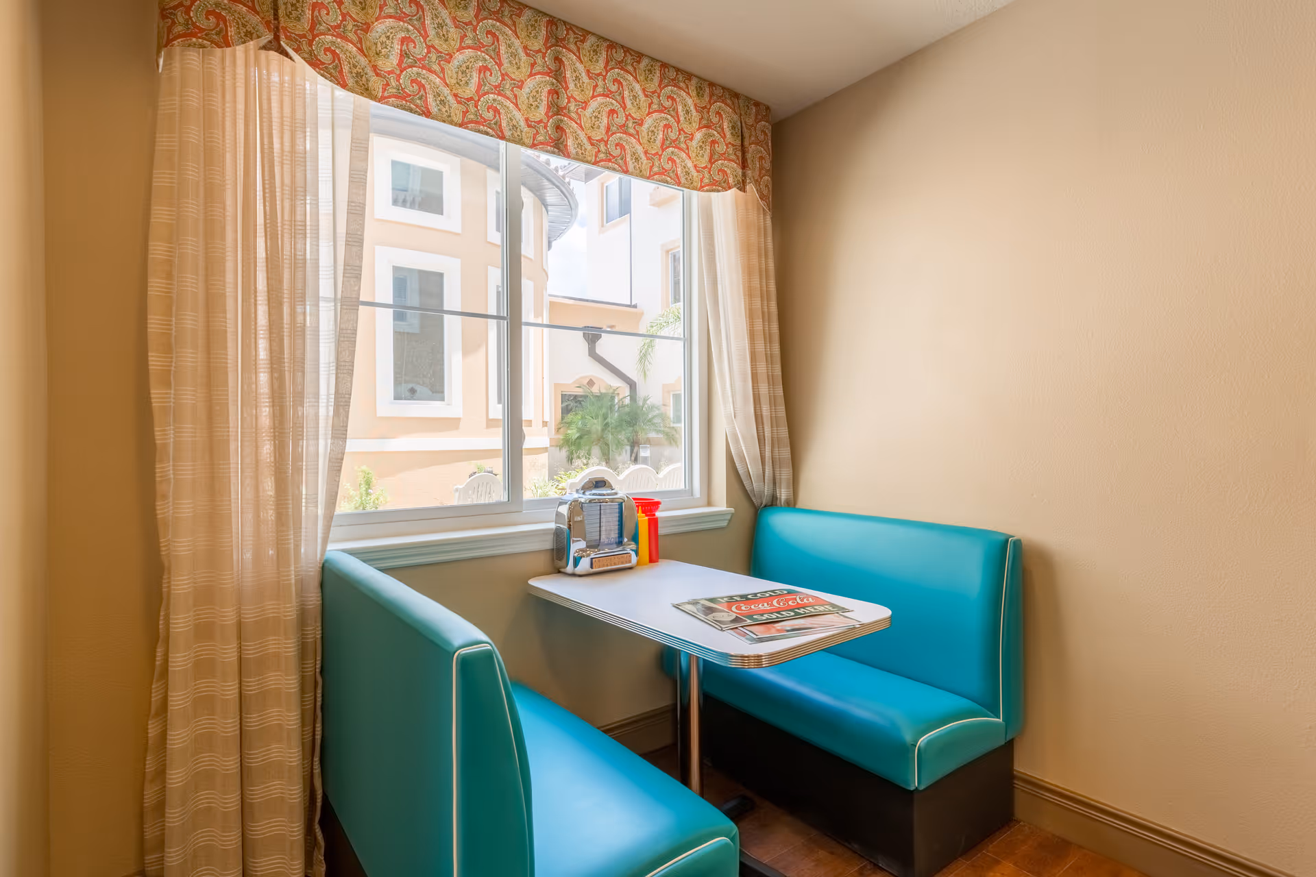 A small dining nook with turquoise upholstered bench seating on two sides of a rectangular table. On the table are a vintage-style napkin dispenser, condiment bottles, and a Coca-Cola menu. The nook is next to a window with sheer curtains and a decorative valance, showing an exterior view of a building with beige walls and white-framed windows.