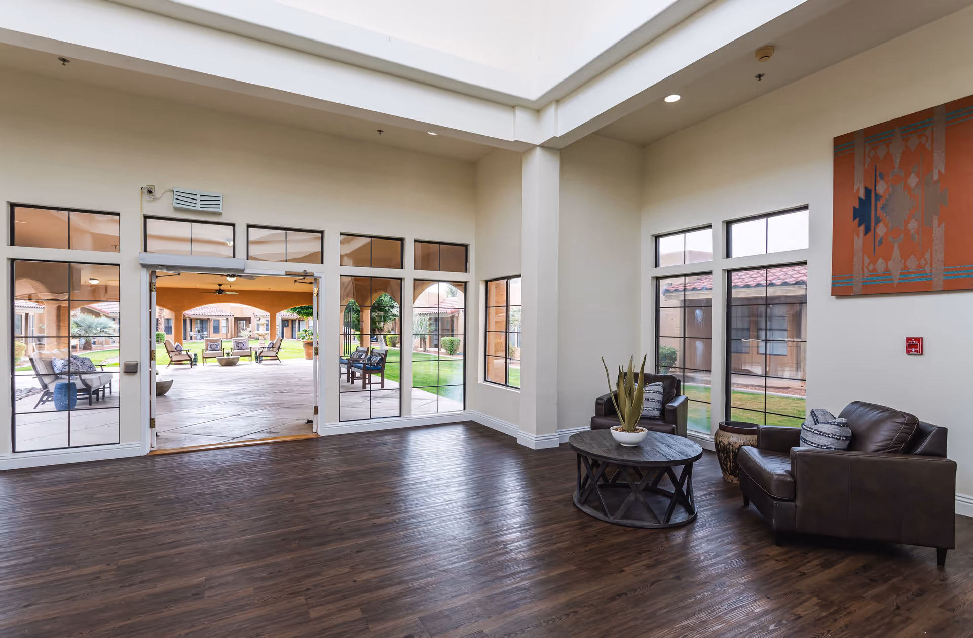 Bright common lounge with leather chairs around a round table, hardwood floors, and large windows/doors opening to an outdoor patio.