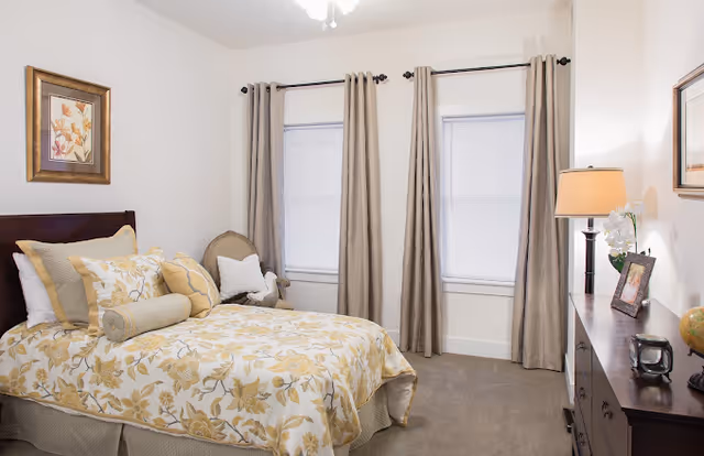 A cozy bedroom with a bed dressed in yellow and white floral bedding, multiple pillows, and a bolster pillow. There are two windows with beige curtains and white blinds. A wooden dresser with a lamp, framed photo, and decorative items is on the right side. A framed floral artwork hangs on the wall above the bed, and a cushioned chair with a white pillow is placed between the windows.