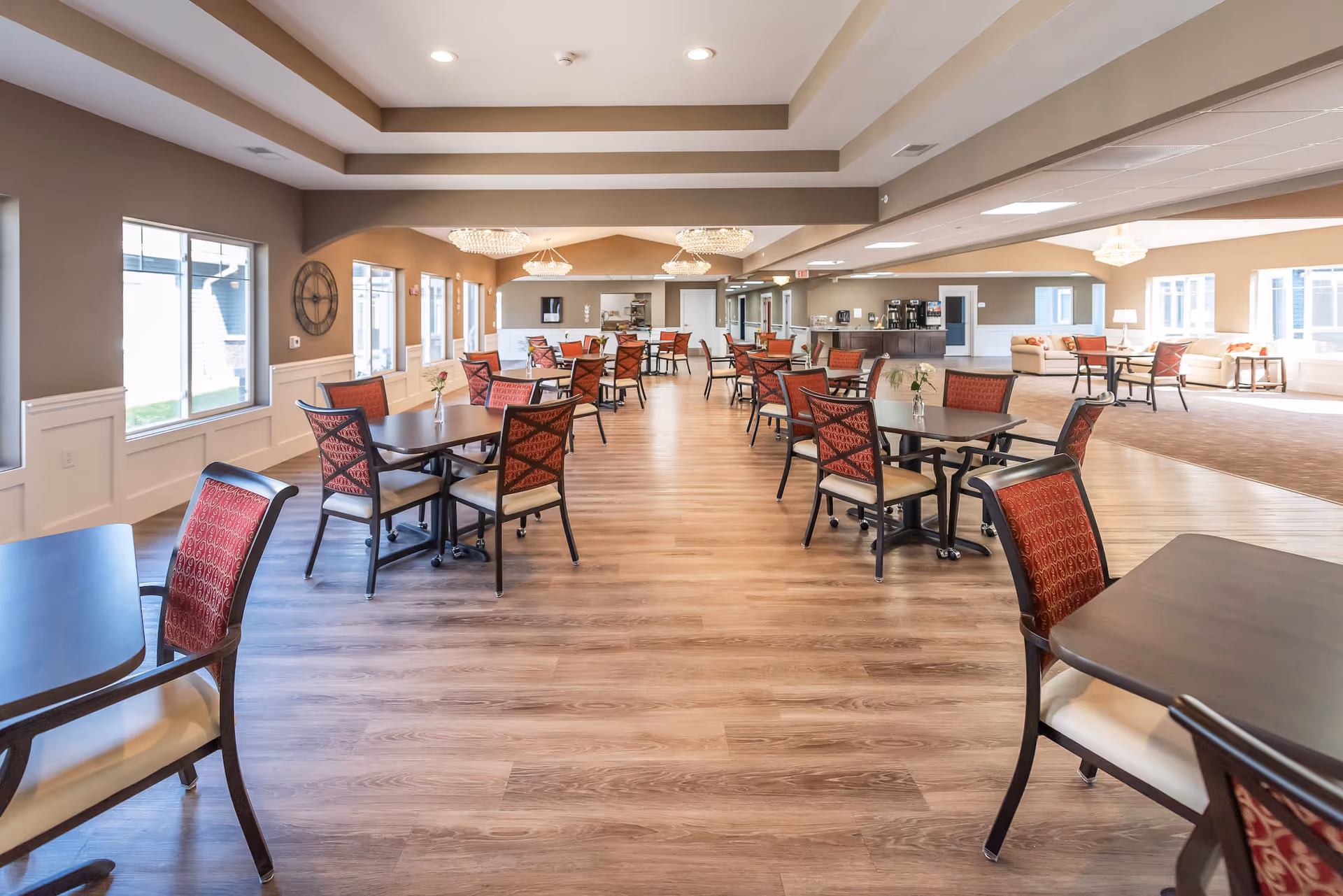 Spacious dining and common area with multiple tables and red-upholstered chairs, wood floors, chandeliers, and lounge seating by the windows.