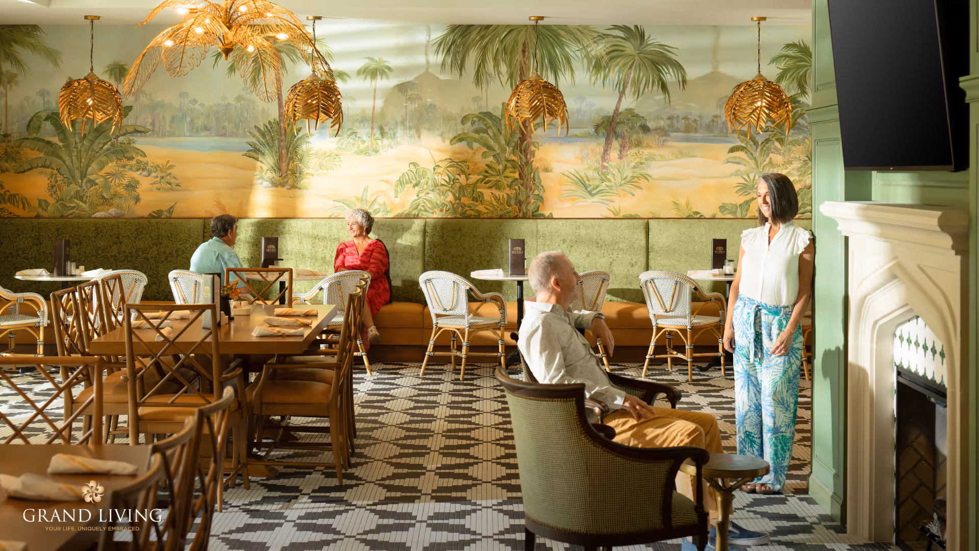 A bright and elegant dining area with patterned tile flooring, wooden tables and chairs, and green cushioned bench seating along the wall. Four seniors are present, two seated and conversing at a table, one man sitting in a chair near a fireplace, and a woman standing nearby. The wall features a large mural of a tropical landscape with palm trees, and there are decorative gold leaf-shaped pendant lights hanging from the ceiling. A television is mounted above the fireplace.