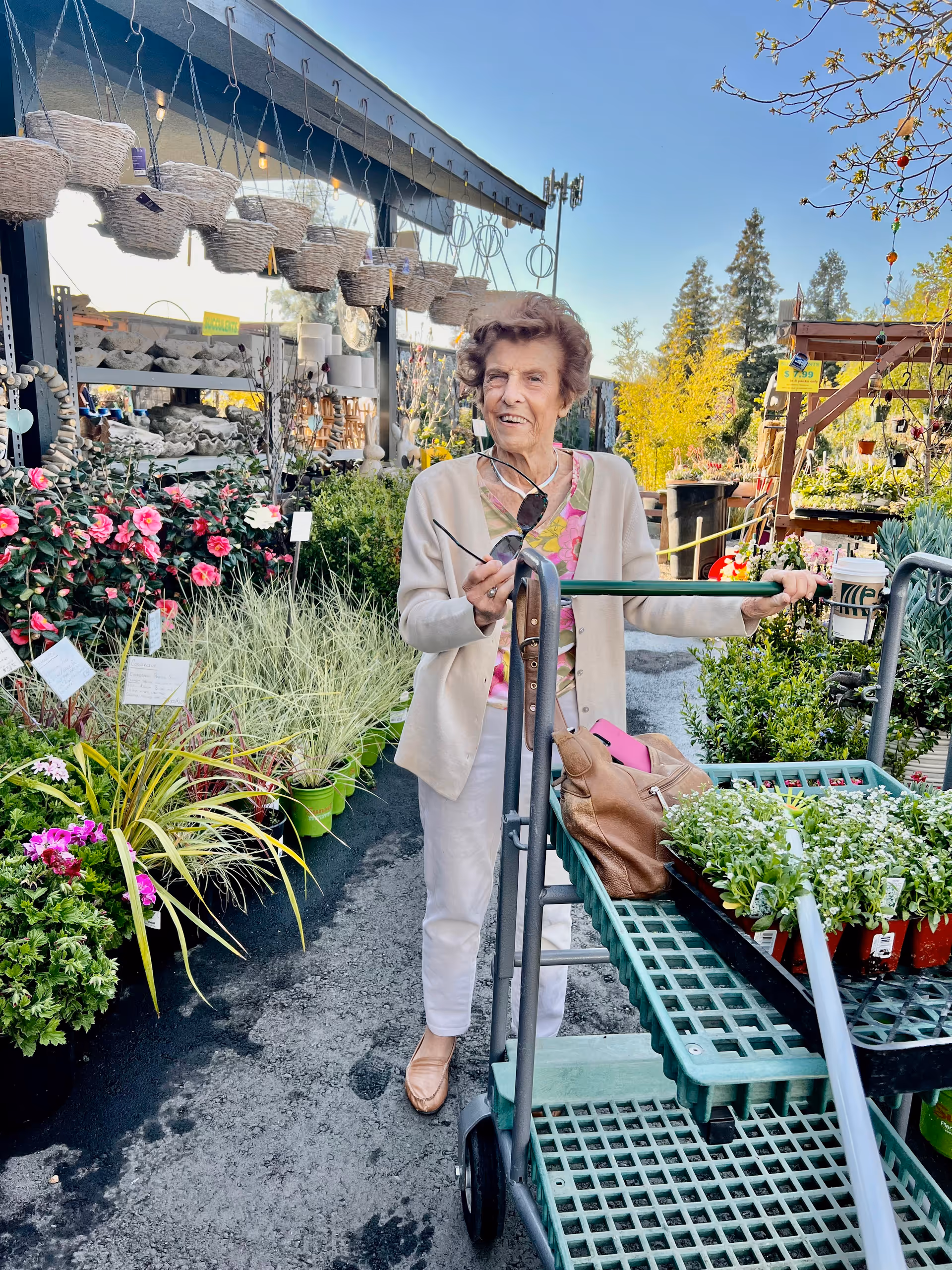 An elderly woman standing outdoors at a garden center, holding a green cart filled with potted plants and a brown handbag. She is wearing a light-colored cardigan, white pants, and tan shoes, and is holding a pair of sunglasses. Surrounding her are various colorful flowers and plants displayed for sale under a clear blue sky.